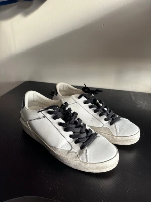 Vita sneakers från Crime med svarta detaljer - Snygga vita sneakers från Crime med svarta skosnören och svart hälparti där loggan syns tydligt. Skorna har en klassisk låg profil och är tillverkade i läder med gummisula. Perfekta för en clean och stilren look.