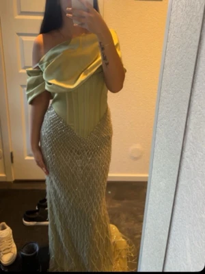 Ljusgrön off shoulder balklänning - Säljer en elegant ljusgrön balklänning med off shoulder-design och draperad överdel. Klänningen har en korsettliknande midja och en lång, figurnära kjol täckt av ett glittrande nätmönster. Perfekt för bal eller festliga tillfällen.