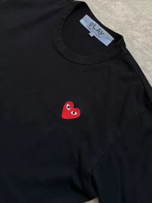 Svart t-shirt märke Cdg - Svart t-shirt med rund hals och ett litet rött hjärta med ögon broderat på bröstet. Enkel och stilren design som passar till det mesta. T-shirten har korta ärmar och är gjord i mjukt bomullstyg.