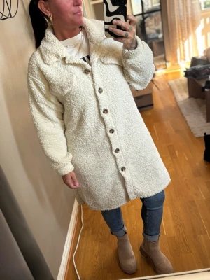 Lång vit teddyjacka med knappar - Säljer en lång, krämvit teddyjacka med stora bruna knappar framtill, inga fickor. Jackan har en oversized passform, mjukt och fluffigt material samt klassisk krage. Perfekt för dig som vill ha en mysig och trendig look.