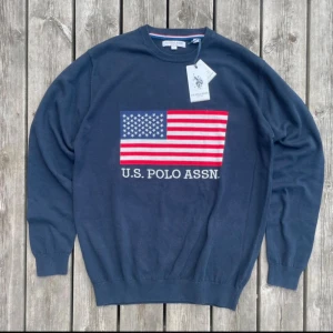 Marinblå tröja U.S. Polo Assn. - Snygg marinblå tröja från U.S. Polo Assn. med stort amerikanskt flaggtryck på bröstet och broderad logga under. Rund halsringning och ribbade muddar. Perfekt för dig som gillar klassisk och sportig stil.