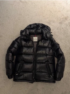 Moncler Maya - Snygg och varm svart dunjacka från Moncler. Jackan har en glansig finish, huva och dragkedja framtill samt två fickor med dragkedja. Perfekt för höst och vinter! Köp direkt eller lägg ett bud.