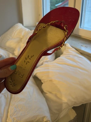 Röda sandaler med guldkedja - Snygga röda sandaler med låg klack och guldfärgad kedjedetalj över foten. Ovandelen är i glansigt material och sulan är beige med dekorativt tryck. Perfekta för att lyfta en outfit med lite extra färg och stil.