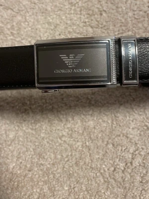 Svart skärp från Giorgio Armani - Snyggt svart skärp från Giorgio Armani med stilren metallspänne där loggan och märket är ingraverat. Skärpet är gjort i slätt skinn och har en klassisk, elegant look som passar till många outfits.