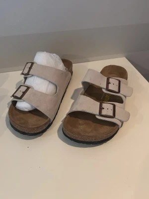 Beige Birkenstock sandaler med spännen - Säljer ett par klassiska Birkenstock sandaler i beige mocka med två breda remmar och justerbara metallspännen. Fotbädden är i brunt läder och sulan är svart. Perfekta för dig som gillar stilren och bekväm design.