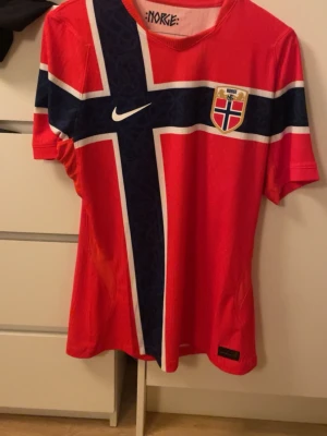 Norge fotbollströja Nike röd M - Säljer en röd Norge fotbollströja från Nike med norska flaggan i blått, vitt och rött över hela framsidan. Tröjan har korta ärmar, rund hals och klubbmärke på bröstet. Perfekt för match eller träning!  Pris kan diskuteras!!
