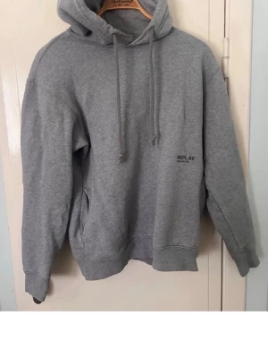 Grå hoodie från Replay - Klassisk grå hoodie från Replay med huva och dragsnören. Tröjan har en enkel design med liten Replay-logga på bröstet och en stor magficka. Tillverkad i mjukt bomullsmaterial och har ribbade muddar vid ärmslut och nederkant.