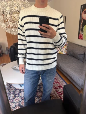 Vit stickad tröja med blåa ränder - Säljer en vit stickad tröja med blåa horisontella ränder. Tröjan har rund halsringning, ribbade muddar och är gjord i ett tjockare material.