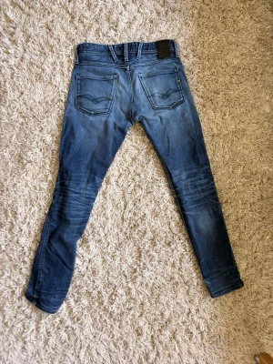 Replay anbass - Säljer ett par blå Replay jeans med snygga slitningar och tvättade detaljer. Jeansen har fem fickor, normal midja och smala ben. Materialet är klassisk denim 30/32