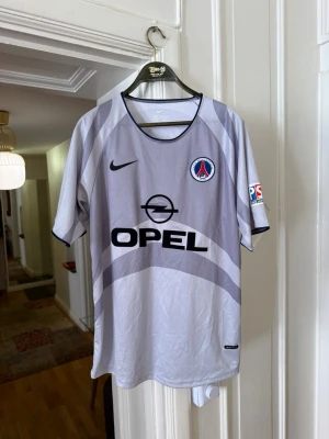 Grå PSG matchtröja från Nike - Säljer en grå PSG matchtröja med svart Nike-logga och Opel som sponsortryck. Tröjan har PSG-emblemet på bröstet och svart kant runt halsen och ärmsluten. Tillverkad i lätt och ventilerande material, perfekt för fotboll eller träning.