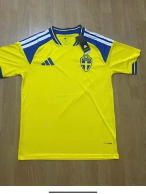 Svensk landslagströja Adidas gul/blå - Säljer en officiell svensk landslagströja från Adidas i gult med blå detaljer och vita ränder på axlarna. Tröjan har Sveriges landslagsemblem på bröstet och är tillverkad i ett lätt, snabbtorkande funktionsmaterial. Perfekt för fotboll eller träning.
