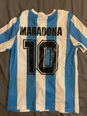 Argentina Maradona tröja Le Coq Sportif - Klassisk Argentina fotbollströja med blå och vita ränder, nummer 10 och Maradona på ryggen. Le Coq Sportif-logga och AFA-märke på bröstet. Perfekt för dig som älskar retro och fotbollshistoria.