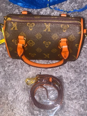Brun handväska med monogram och orange detaljer - Snygg brun handväska med klassiskt monogrammönster och orange handtag samt detaljer. Väskan har guldfärgade metalldelar och medföljande axelrem. Perfekt storlek för det viktigaste och ger en lyxig känsla till din outfit.