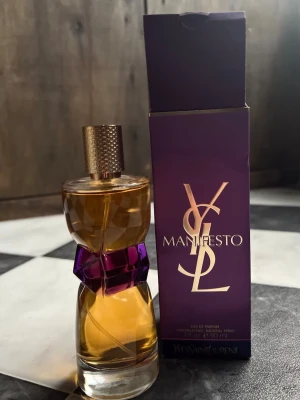 YSL Manifesto EdP 90 ml discontinued  - YSL Manifesto Eau de Parfum i en elegant glasflaska med lila och guld detaljer. Flaskan har en unik form med en lila midja och guldigt lock. Parfymen kommer i originalförpackning med lila och guld design. Innehåller 90 ml. Den är bara testad och är som ny. Lådan är något skadad som syns på bilderna. 