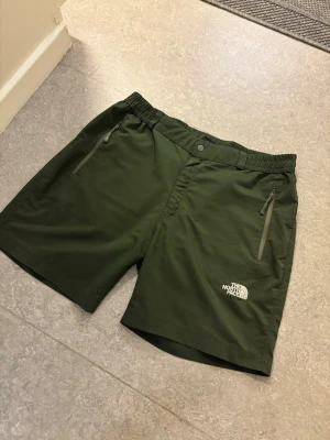 Gröna shorts från The North Face - Mörkgröna shorts från The North Face med elastisk midja och dragkedjefickor. Perfekta för sommarens äventyr eller träning. Lätt och smidigt material med logga broderad på ena benet. Snygg och sportig look.
