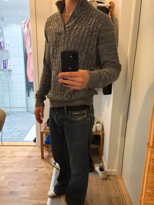 Grå stickad tröja  - Säljer en grå stickad tröja med knappar och hög krage. Tröjan har ett snyggt kabelstickat mönster framtill och ribbade muddar vid ärmslut och nederkant. Perfekt för kyliga dagar och enkel att matcha med jeans.