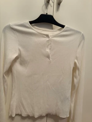 Vit ribbad långärmad topp - Snygg vit ribbad topp med lång ärm och rund halsringning. Toppen har knappar framtill och är tillverkad i ett stretchigt material som sitter skönt på kroppen. Perfekt att styla med jeans eller kjol för en enkel och fräsch look.