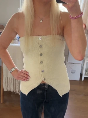 Beige stickad tubtopp med knappar - Säljer en beige stickad tubtopp med dekorativa knappar framtill. Toppen är axelbandslös och har en unik formad nederkant som ger en cool look. Perfekt att styla med jeans eller kjol för en trendig outfit.