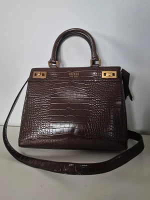 Guess Katey Croc Luxury Satchel handväska 30 cm - Handväska från Guess (modell: Katey Croc Luxury Satchel).   Mycket rymlig och välorganiserad modell (bredd ca 30 cm) med flera smarta fack på insidan samt ett praktiskt blixtlåsfack på baksidan.  Jättefint skick! Mycket sparsamt använd, ren och fräsch både på utsidan och i det logomönstrade innerfodret (se bilder).      Nypris: 1 356 kr. 