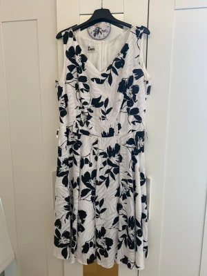 Vit klänning med svart blommönster - Snygg ärmlös klänning i vitt med svart blommigt mönster och v-ringning. Klänningen har en markerad midja och ett utsvängt kjolparti som ger en feminin siluett. Klänningen har också en dragkedja på baksidan. Perfekt för sommarens alla tillfällen.                                           Är öppen för att förhandla priset🌸