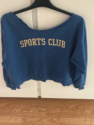 Blå croppad sweatshirt SPORTS CLUB - Blå croppad sweatshirt med texten 'SPORTS CLUB' i beige på bröstet. Tröjan har en vid, rå kantad halsringning och långa ärmar med muddar. Perfekt för en avslappnad och sportig look.