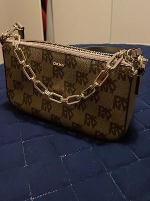 Brun monogram handväska från DKNY - Snygg brun handväska från DKNY med monogramtryck och silverfärgad kedja. Väskan har justerbar axelrem i läderimitation och dragkedja upptill. Perfekt storlek för det viktigaste och ger en trendig look till din outfit.(den är äkta)