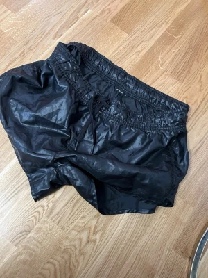 Svarta träningsshorts från SOC - Säljer ett par svarta träningsshorts från SOC i storlek 40. Shortsen har elastisk midja med snörning och är gjorda i ett glansigt syntetmaterial som känns lätt och smidigt. Perfekta för gymmet eller löprundan!