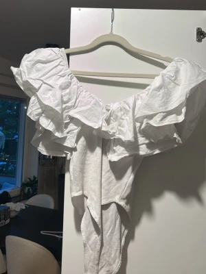 Vit volang body från Zara - Säljer en superfin vit topp från Zara med stora volanger över axlarna. Toppen är axelbandslös och har en offshoulder-design som ger en trendig look. Perfekt för dig som vill sticka ut lite extra med en unik och luftig stil.