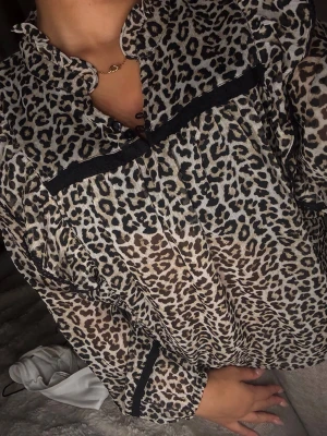 Leopardmönstrad volangblus - Trendig blus med leopardmönster i beige, svart och brunt. Blusen har volangdetaljer längs axlarna, svarta band och en liten ståkrage. Tillverkad i ett tunt, lätt transparent material och har långa ärmar för en cool och modern stil.