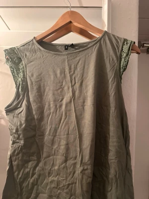 Grön ärmlös topp med spetsdetalj - Snygg grön ärmlös topp med spetsdetaljer vid axlarna. Toppen har rund halsringning och är tillverkad i ett mjukt bomullsmaterial. Perfekt för en avslappnad och trendig look.