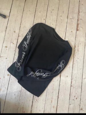 Forever faithful sleeve  - Svart långärmad tröja med vit broderad text 'Forever Faithful' längs båda ärmarna. Tröjan har rund halsringning och är gjord i ett mjukt, strukturerat material. Perfekt för en stilren och avslappnad look.
