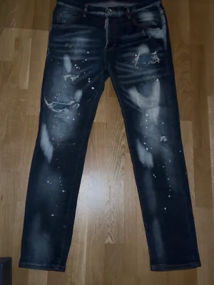Mörkblå dsquared - Säljer ett par mörkblå jeans med slitningar, hål och vita färgstänk för en riktigt edgy look. Jeansen har raka ben, fem fickor och coola detaljer både fram och bak. Perfekta för dig som gillar streetwear och vill sticka ut.