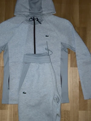 (Helt ny) Grå Lacoste tracksuit, tröja M, byxor S - Snygga gråa mjukisbyxor från Lacoste med klassisk krokodillogga på benet. Byxorna har resår i midjan och sidofickor. Perfekta för en avslappnad och sportig stil. Matchar med hoodie i samma färg och material.