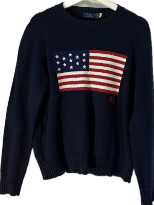 Ralph Lauren USA tröja  - Mörkblå stickad tröja från Polo Ralph Lauren med en stor amerikansk flagga i rött, vitt och blått på bröstet. Tröjan har rund halsringning, ribbade muddar och RL-broderi. Perfekt för dig som vill ha en klassisk och stilren look. ALDRIG ANVÄND 