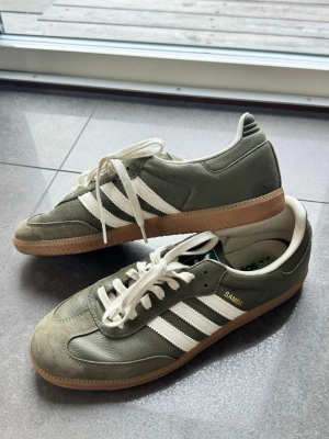 Adidas Samba gröna sneakers - Säljer ett par klassiska Adidas Samba sneakers i grönt läder med vita detaljer och gummisula. Skorna har de ikoniska tre ränderna på sidan och texten 'SAMBA' i guld. Perfekta för dig som gillar retrostil och streetwear.