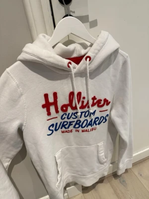 Vit Hollister hoodie - Snygg vit hoodie från Hollister med broderat rött och blått tryck på bröstet där det står 'Hollister Custom Surfboards'. Hoodien har huva med dragsko, magficka och mjukt material på insidan. Perfekt för en avslappnad och cool stil. Rätt så sällsynt då dom endast gjordes under 2000,,,