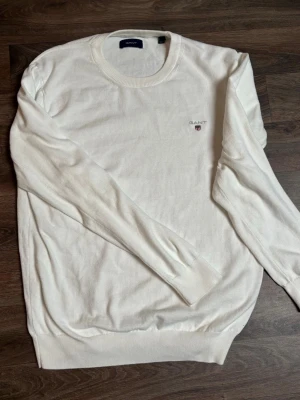 Vit stickad tröja från GANT - Klassisk vit stickad tröja från GANT med rund halsringning och diskret logga broderad på bröstet. Tröjan har långa ärmar och ribbade muddar vid ärmslut och nederkant. Perfekt för en clean och stilren look.