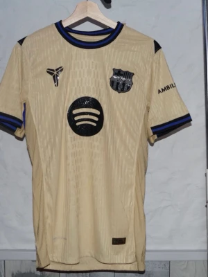 Beige Barcelona matchtröja - Snygg beige Barcelona matchtröja med svarta detaljer och klubbmärke på bröstet. Tröjan har svarta och blå ränder på krage och ärmslut samt tryckta loggor på bröstet och ärmen. Tillverkad i lätt och ventilerande material, perfekt för fotboll eller träning.
