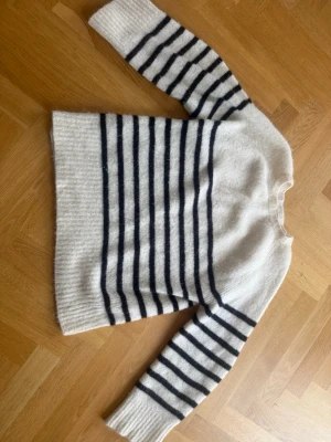 Vit stickad tröja med svarta ränder - Mysig stickad tröja i vitt med svarta horisontella ränder på både kropp och ärmar. Tröjan har rund halsringning, lång ärm och ribbstickade muddar. Perfekt för dig som gillar klassisk och avslappnad stil.