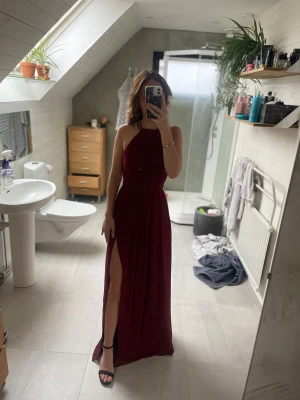 Vinröd balklänning med strassdetaljer  - Hej! Säljer min balklänning då den inte längre kommer till användning. Det är en lång klänning med slits vid benet och pärlor och diamanter längst med ryggen. Jag är 162 cm lång men skulle säga att klänningen kan bäras av alla mellan 160-170 cm beroende på hur höga klackar man har😊 klänningen är nytvättad och har inga fläckar eller skavanker!