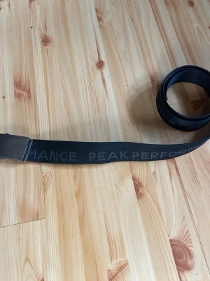 Svart bälte från Peak Performance - Snyggt svart bälte från Peak Performance med diskret logga längs hela bandet. Bältet har en metallspänne och är gjort i slitstarkt material som passar till jeans eller byxor. Perfekt accessoar för en clean och sportig look.