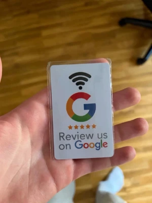 Google NFC Recensionstag - En liten rektangulär NFC-tagg i plast med Google-logga och texten 'Review us on Google'. Perfekt för företag som vill göra det enkelt för kunder att lämna recensioner genom att scanna taggen med mobilen. Vit bakgrund med färgglad logga och svart wifi-symbol.