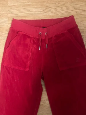 Röda mjukisbyxor i velour - Snygga röda mjukisbyxor i mjuk velour med bred resår i midjan och dragsko. Byxorna har stora fickor fram och en bakficka med knappdetalj. Perfekta för en chill och avslappnad stil.