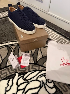 Mörkblå christian louboutins - Snygga mörkblå sneakers från Christian Louboutin i mocka med matchande nitar på tån och vita sulor. Skorna har klassisk låg profil, ton-i-ton snörning och exklusiv känsla. Perfekta för dig som vill sticka ut med en lyxig touch.