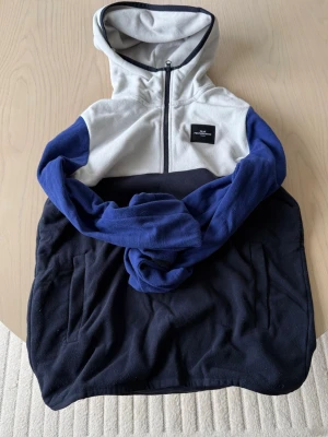 Fleecehoodie från Peak Performance  - Snygg hoodie från Peak Performance i fleece med färgblock i blått, svart och ljusgrått. Tröjan har dragkedja framtill, huva och två fickor. Patch på bröstet. Storlek S. Bra skick.
