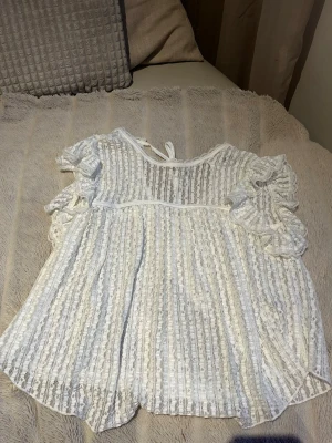 Vit spetsblus med volangärmar - Söt vit blus i spets med korta volangärmar och rund hals. Blusen har dekorativa band i nacken och ett luftigt, romantiskt fall. Perfekt för dig som gillar en feminin och trendig stil. Materialet är genombruten spets med blommönster.