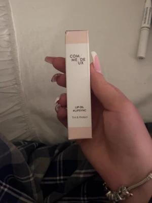 Comme Deux Lip Oil #LIPSYNC - Säljer ett Comme Deux Lip Oil #LIPSYNC som ger både färg och skydd till läpparna. Förpackningen är stilren i beige och brunt, och själva läppoljan kommer i en smidig tub med skruvlock. Perfekt för dig som vill ha återfuktade och snygga läppar.