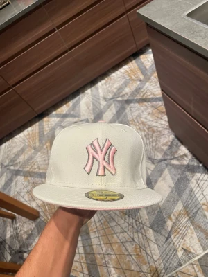 Vit New York Yankees keps med brodyr - Snygg vit keps från New Era med New York Yankees-logga i ljusrosa framtill. På sidan finns en detaljerad brodyr med blommor och grått motiv. Klassisk flat peak och justerbar passform. Perfekt accessoar för dig som vill sticka ut.