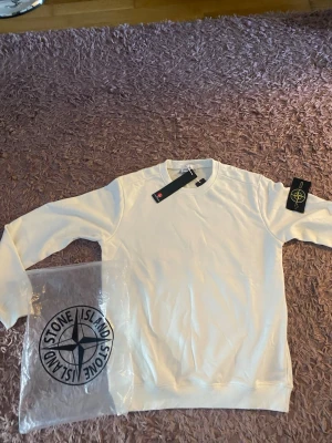 Vit sweatshirt från Stone Island - Snygg vit sweatshirt från Stone Island med klassisk rund hals och ribbade muddar. Tröjan har en stilren design utan tryck och kommer med originaltaggar och påse. Perfekt för dig som gillar streetwear och enkel stil.