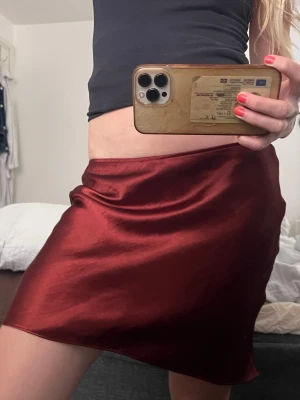 Vinröd satin kortkjol - Säljer en vinröd kortkjol i glansigt satinmaterial. Kjolen har en enkel och stilren design med lätt utsvängd form och slät midja. Ungefär i kort- midi längd. 